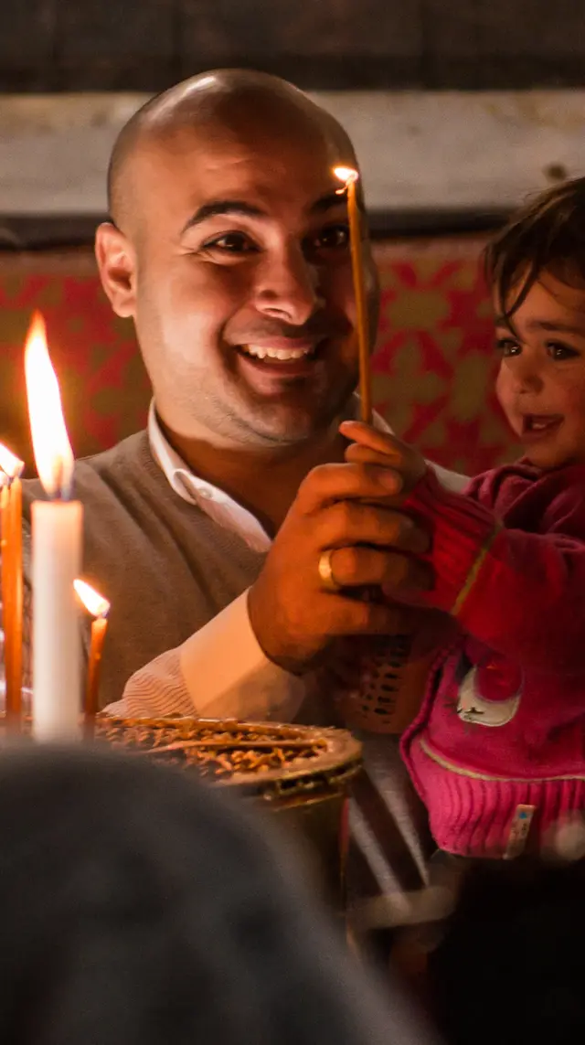 A father holding his daughter as she reaches for the candle he has just lit.