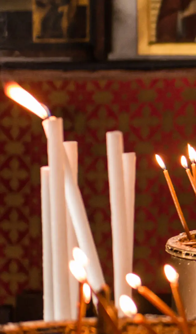 Thin beeswax tapers and tall white candles burning in a stand before the red damask wall.